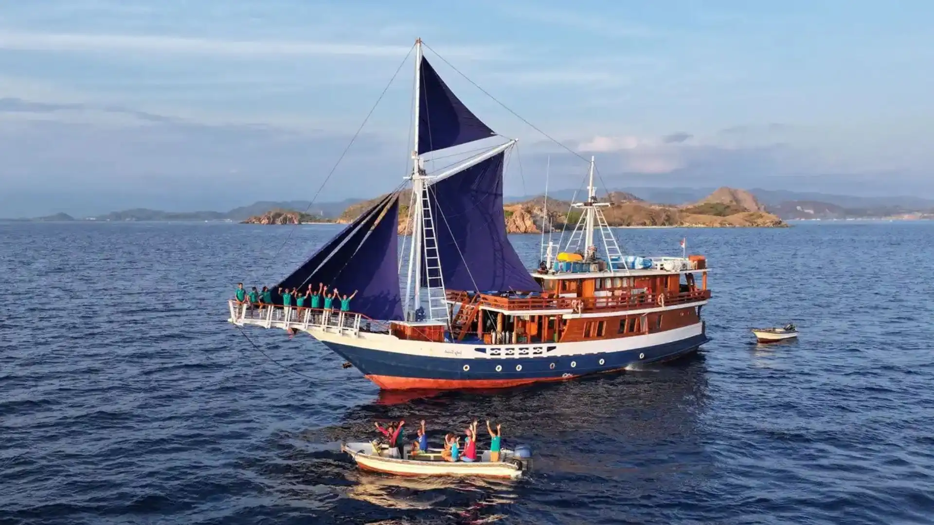 Lady Denok Liveaboard sailing with guests on the bowsprit and dive tenders alongside | Komodo Luxury